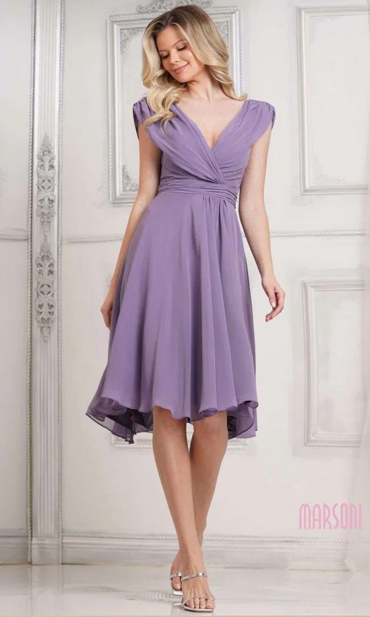 A woman stands indoors against a light decorative wall, wearing the Marsoni M251S short semi-formal dress in wisteria, featuring lilac chiffon fabric, knee-length hem, deep v-neckline, and cap sleeves.